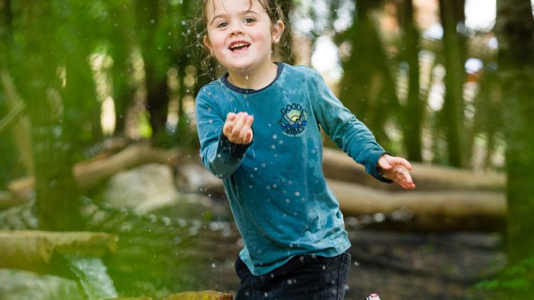 Young girl in teal t-shirt and black shorts splashing water on the camera whilst playing in our Nature's Playground natural outdoor play area