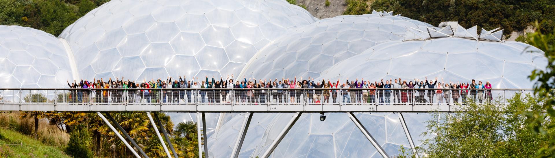 People standing on a bridge in front of the Eden Project Biomes with their hands in the air