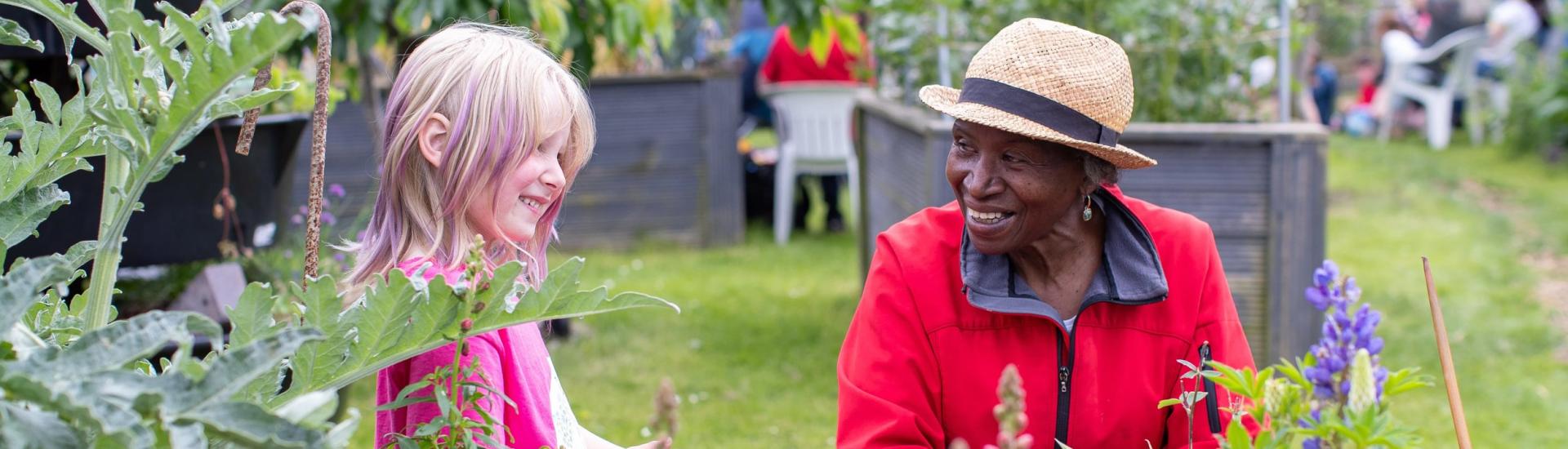 An older woman and younger girl smile at each other in a colourful community garden